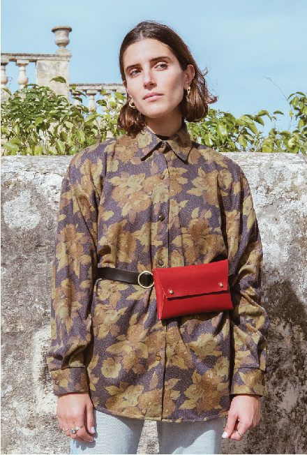 A woman wears a vintage charcoal, floral shirt with a red  leather belt bag around her waist.