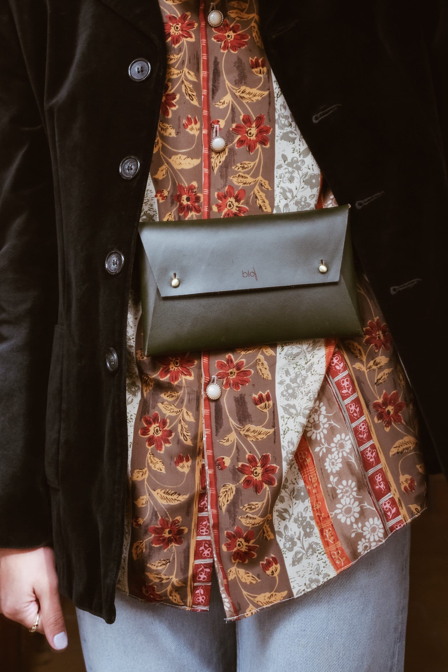 A woman wears a paisley vintage shirt with a black velvet jacket and a green leather belt bag around her waist. 